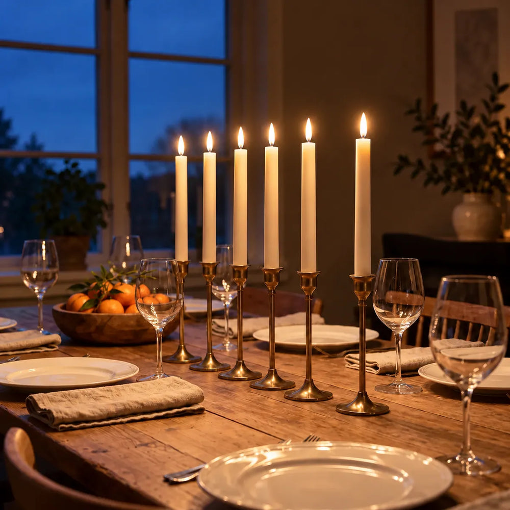 Six lit Wove Luma LED taper candles in brass holders on a laid dining table on a winter evening