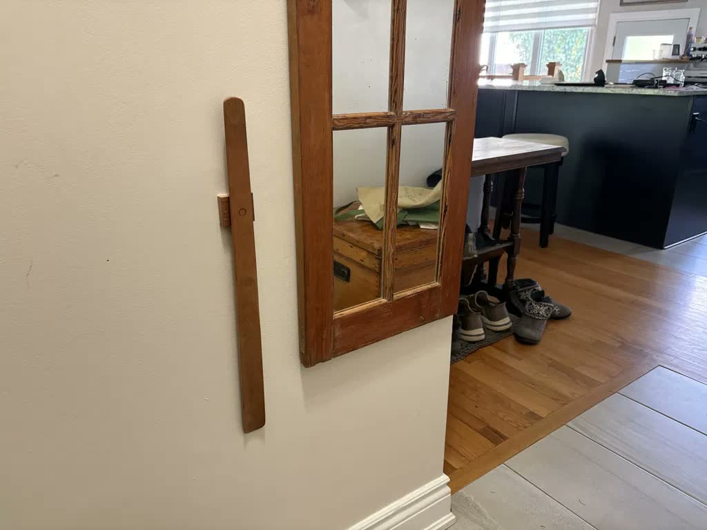 Light oak shoehorn on a cream wall beside an old raw-wood-framed mirror, with a view into the kitchen