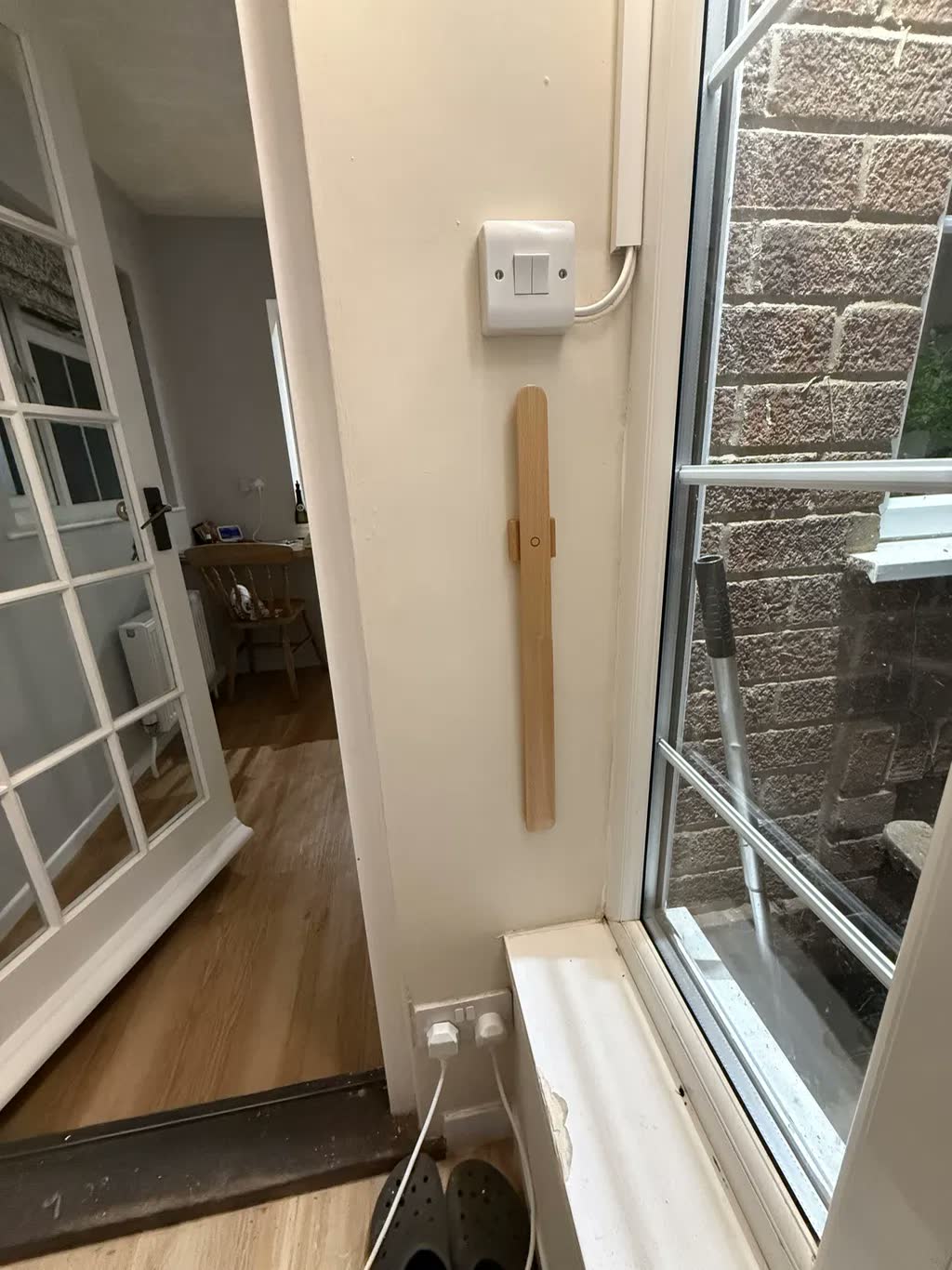 Light oak shoehorn on a cream wall by a window and doorway, with a light switch above and clogs on the floor