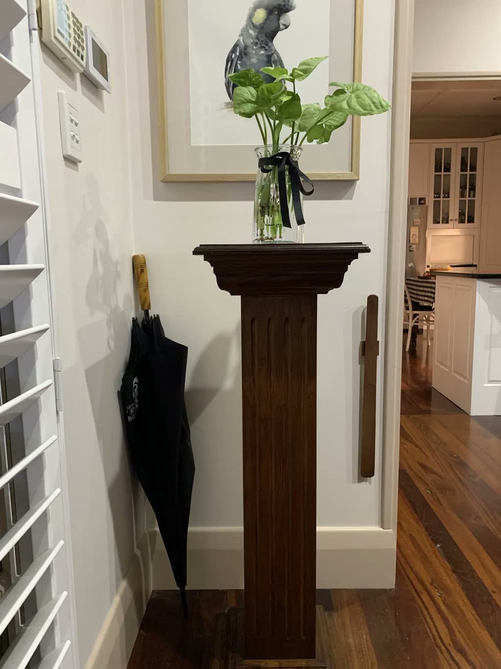 Dark walnut shoehorn on a white hallway wall beside a dark antique pedestal table with a glass vase and a black umbrella